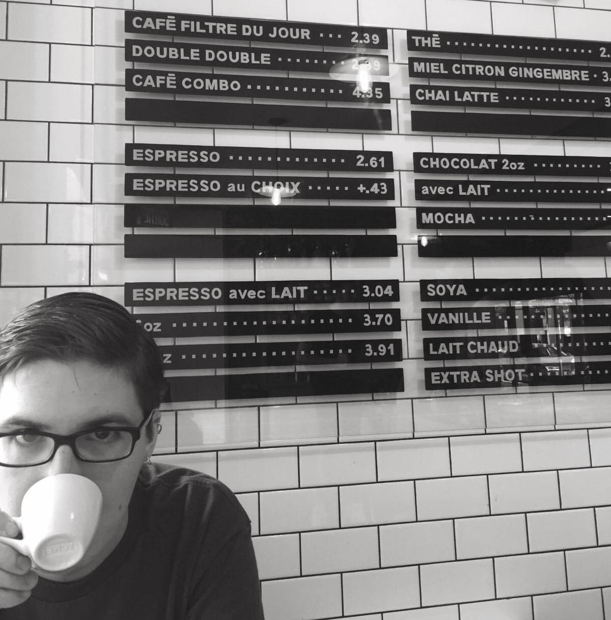 person sitting in a cafe drinking an espresso and staring at the camera in the bottom left of the image, behind on the wall is a menu depicting various coffee drinks