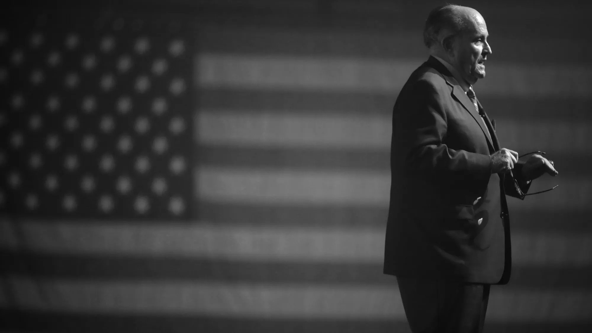 Rudy Giuliani standing on the left of the image looking away to the right, with an American flag out of focus behind him