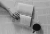 A hand holds a book open on a bed of book pages next to a cup of coffee, seen from above.